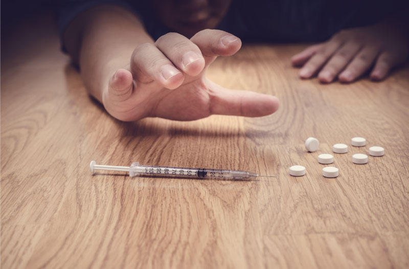 person with drug addiction reaching for needle and pills in foreground